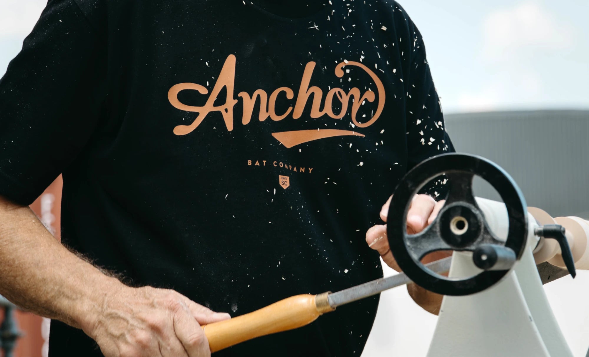 A bat maker at the lathe in an Anchor Bat Company tee, wood shavings in the air